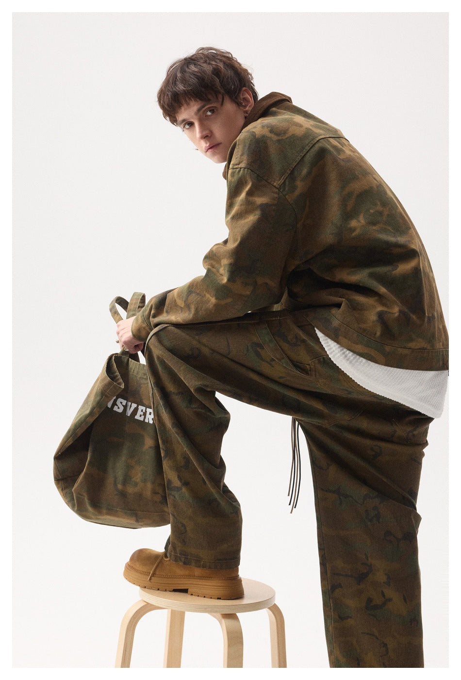 Model seated sideways in a brown-and-olive camouflage jacket and matching loose trousers, showing the jacket's back and side.