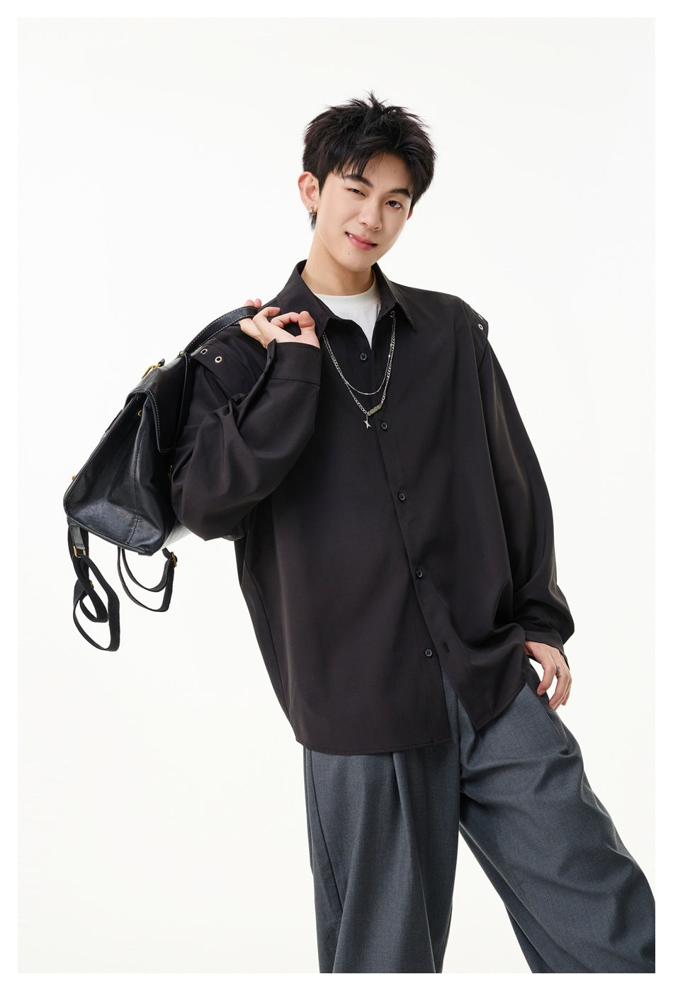 Model wearing an oversized black button-up shirt with shoulder snaps and silver chains, holding a black bag.