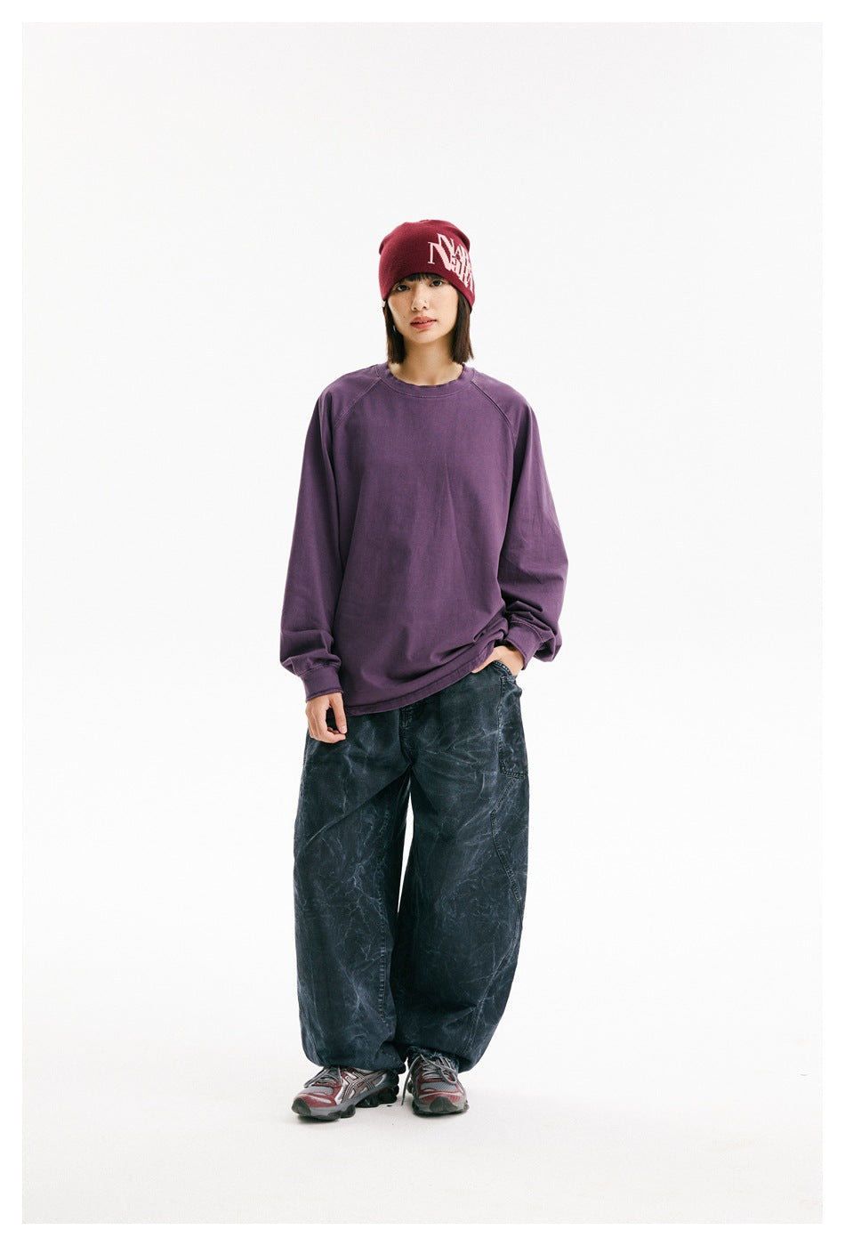 Model in a purple oversized raglan sweatshirt, dark patterned loose pants, and a burgundy beanie with white lettering.
