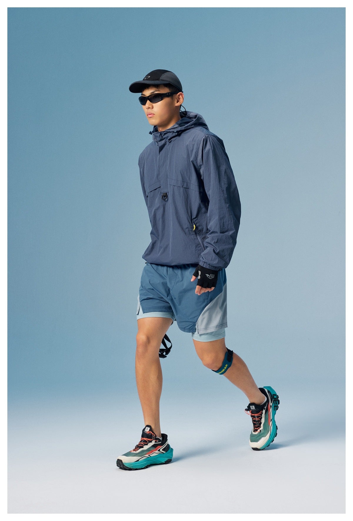 Model wearing a slate-blue hooded pullover jacket and blue-gray running shorts outdoors-style against a blue studio backdrop.