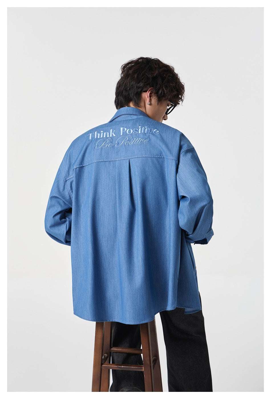 Back view of a person seated on a wooden stool wearing a blue shirt with embroidered Think Positive Be Positive script