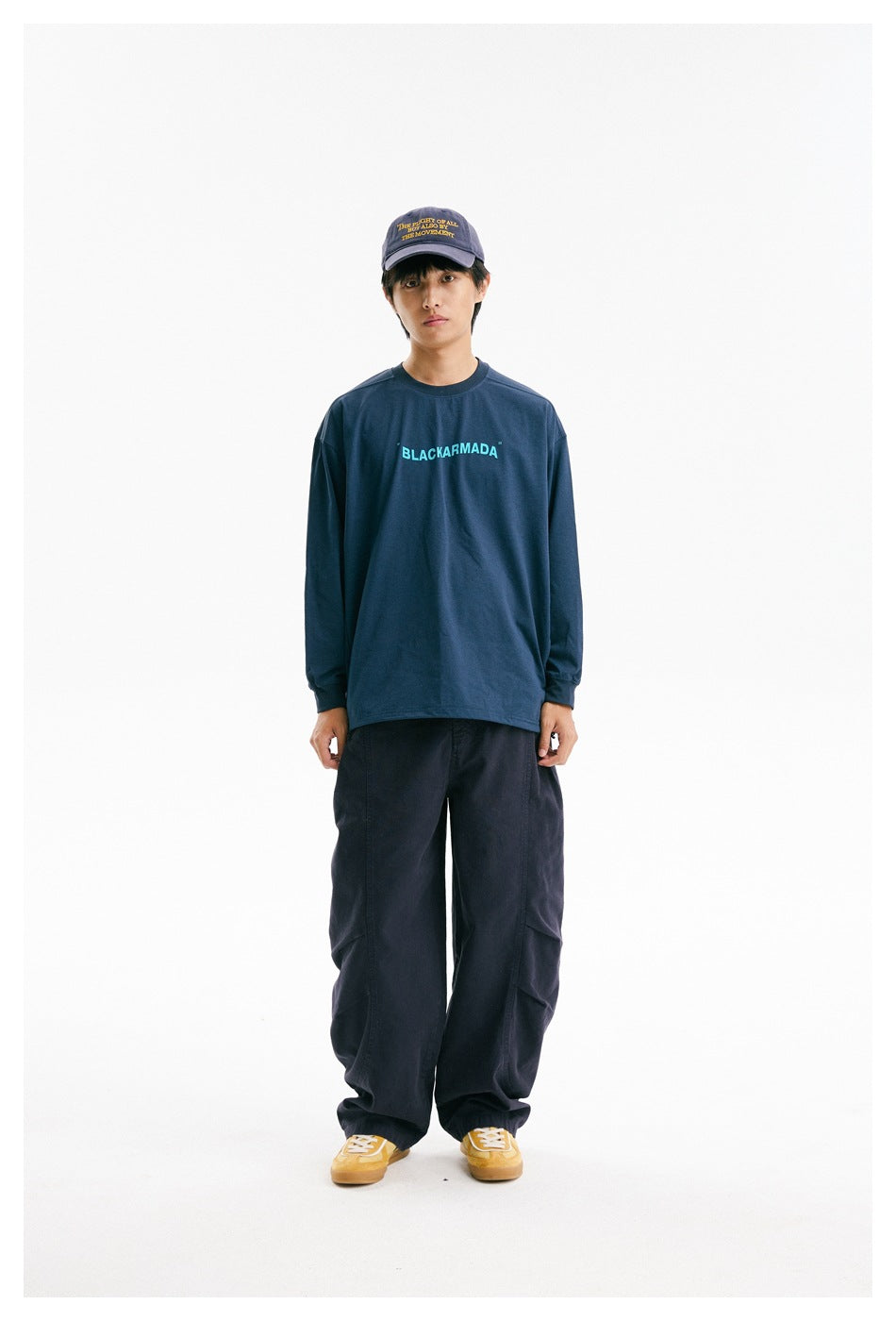 Model wearing a navy long-sleeve shirt with cyan chest lettering, loose dark pants, and a purple cap.