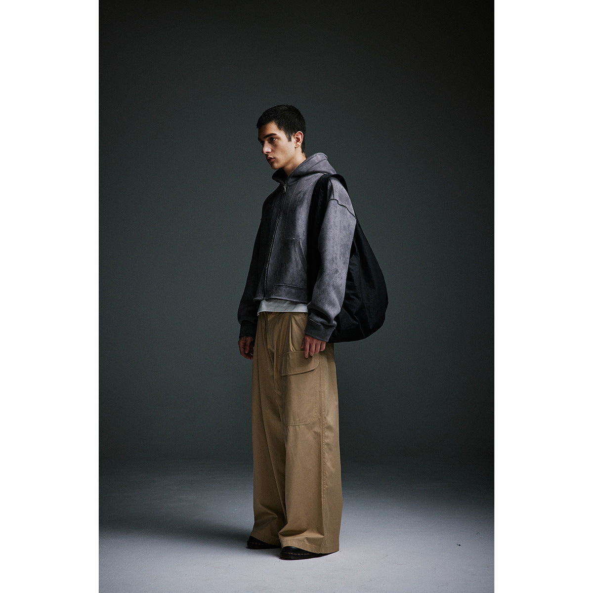 Model in a charcoal zip-up hoodie, loose taupe cargo trousers and a black shoulder bag, shown in profile.