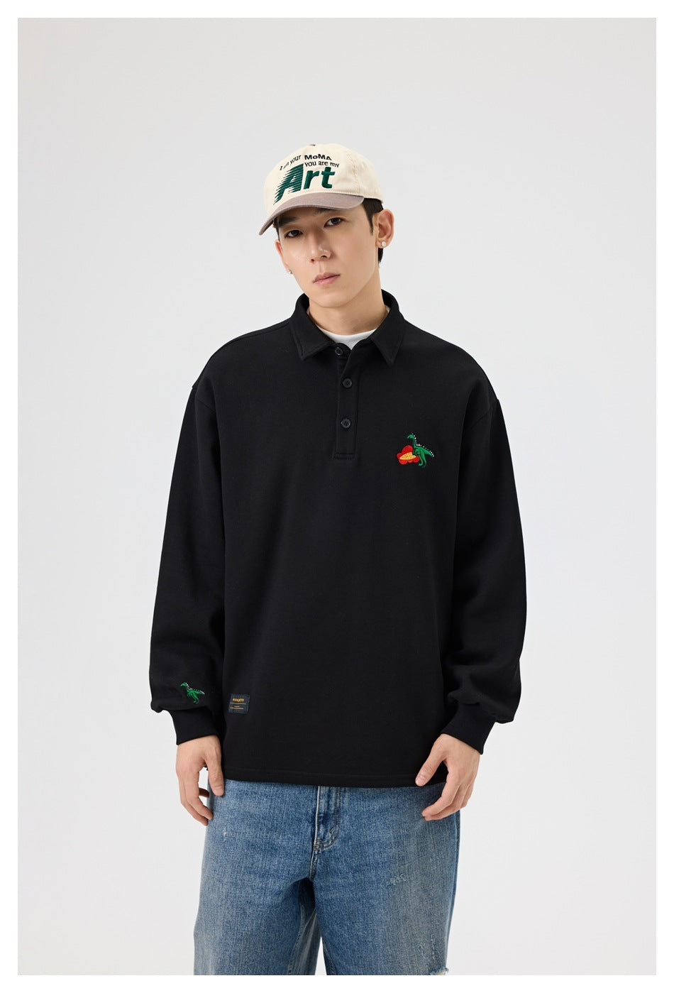 Model wearing black polo-collar top with embroidered dinosaur, cap and light-wash jeans, front view