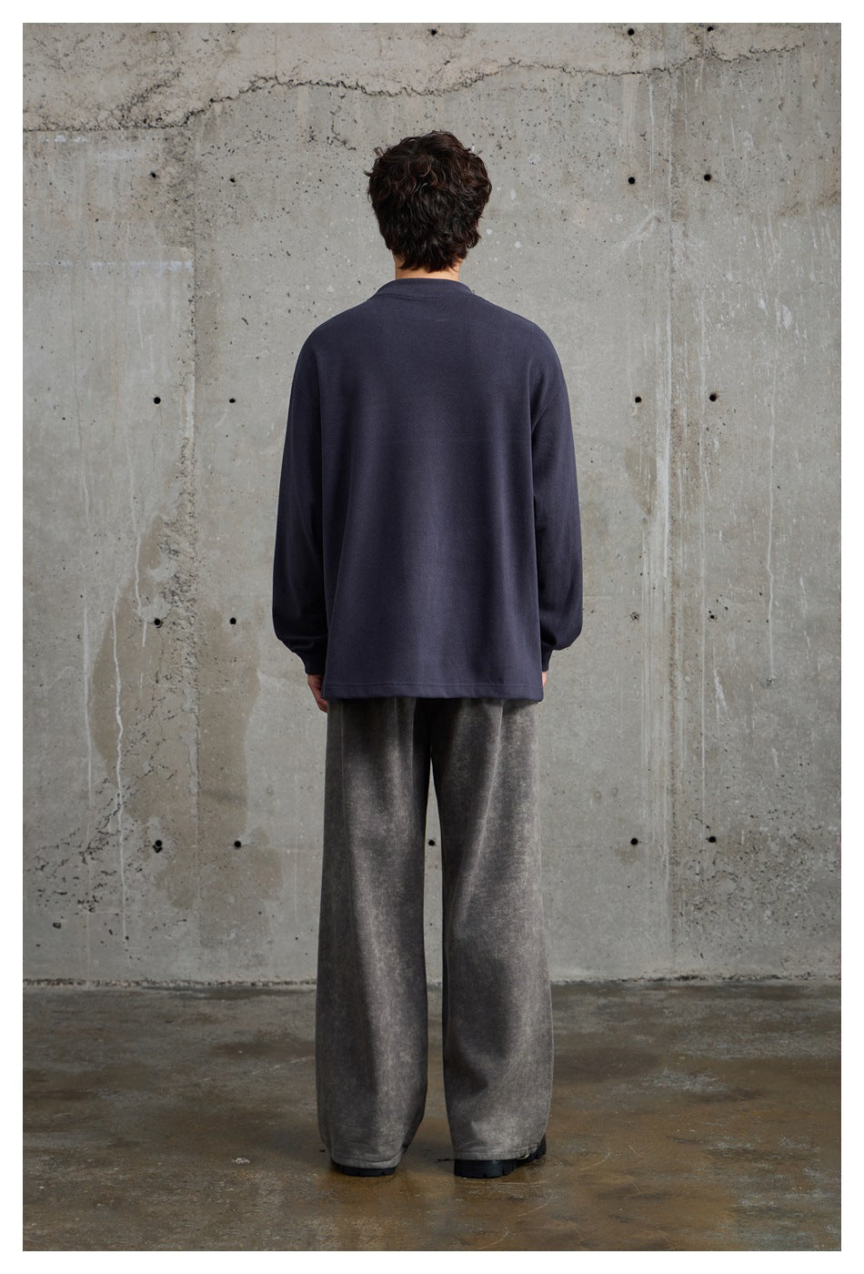 Rear view of a model wearing a plain navy oversized crewneck and loose gray trousers.