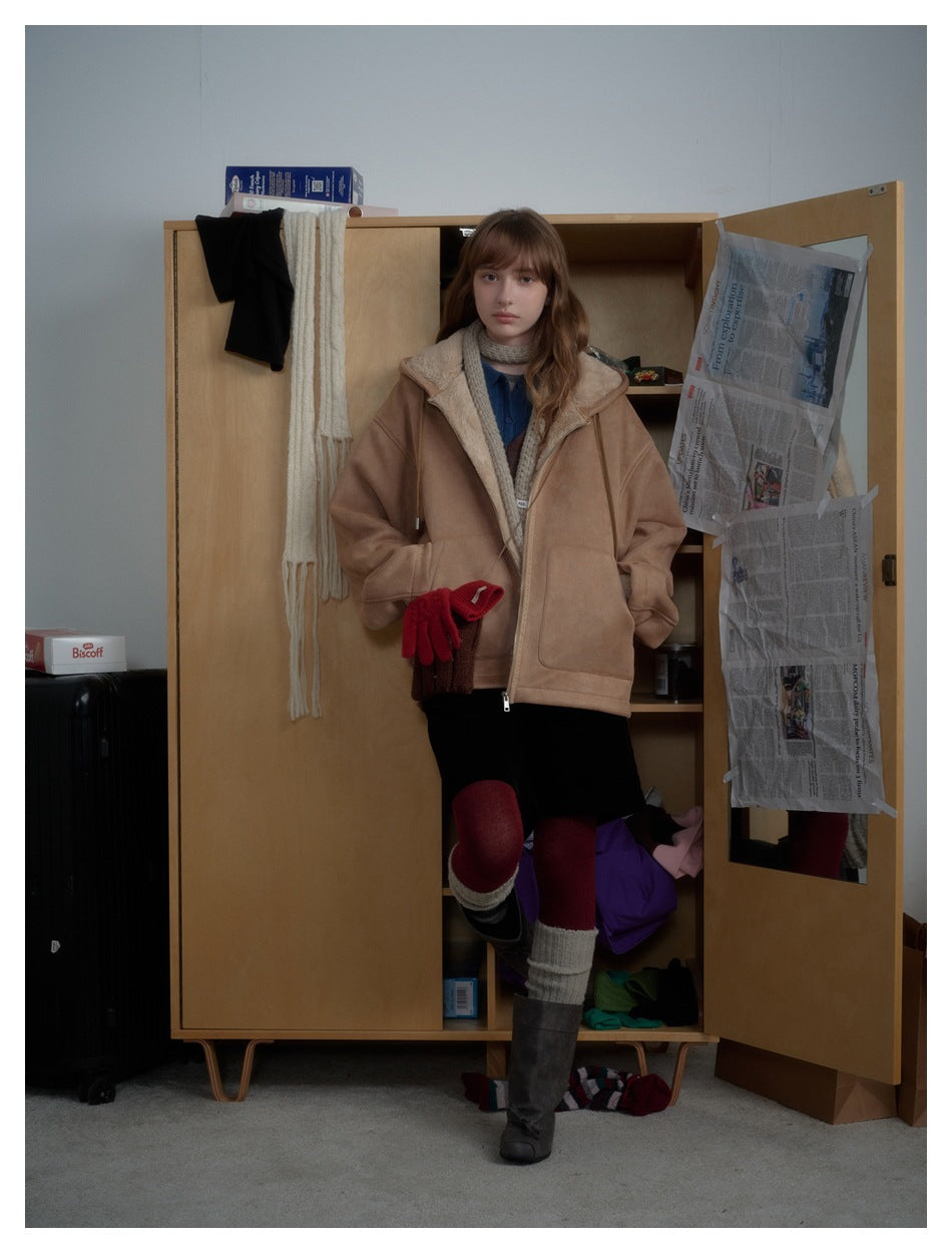 Model wearing the brown hooded zip jacket open over a scarf and dark skirt, hand in pocket, standing by a wooden wardrobe