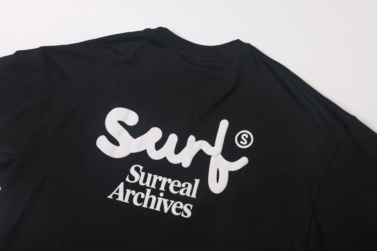 Back of a black short-sleeve shirt with a large white stylized logo and “Surreal Archives” text.