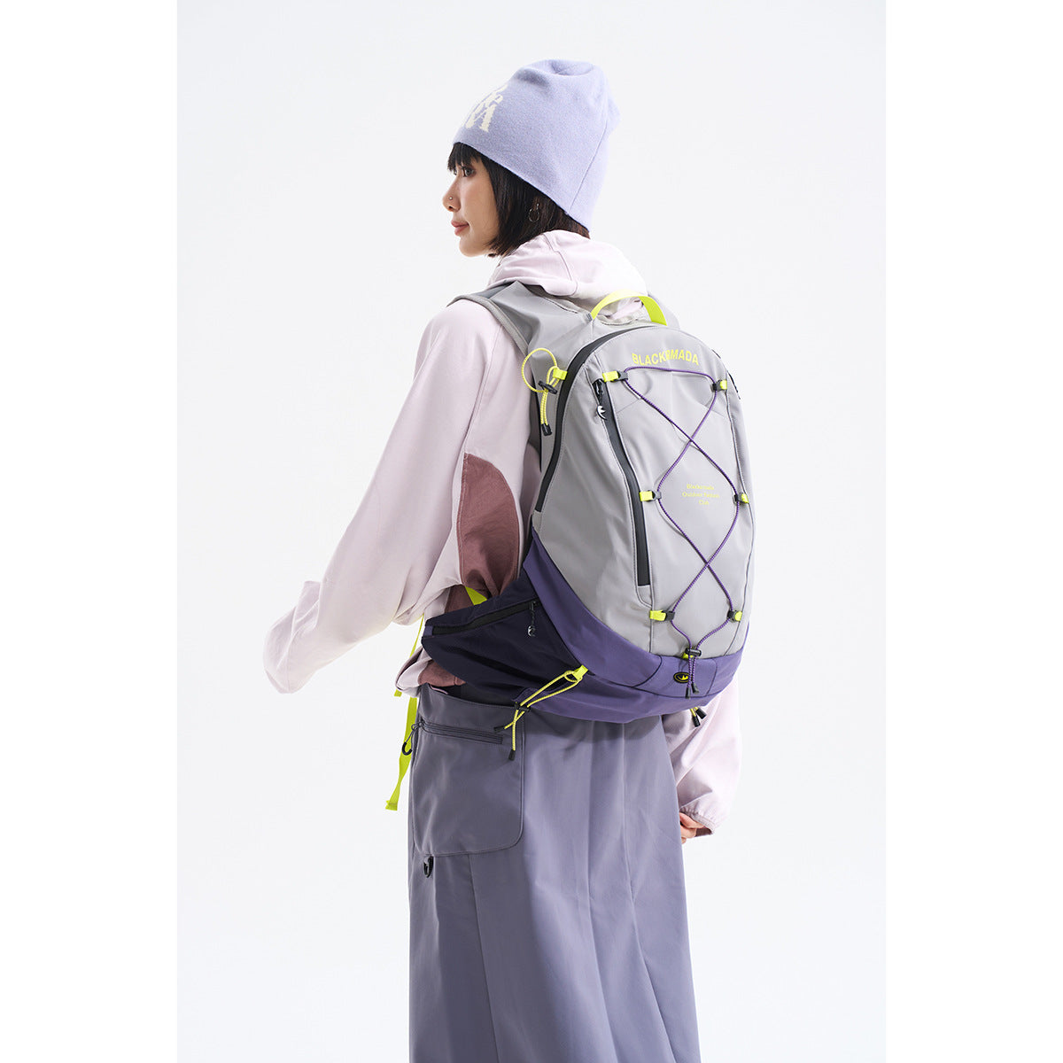 Model seen from behind wearing a gray and purple hiking backpack with yellow accents and crossed elastic cord.
