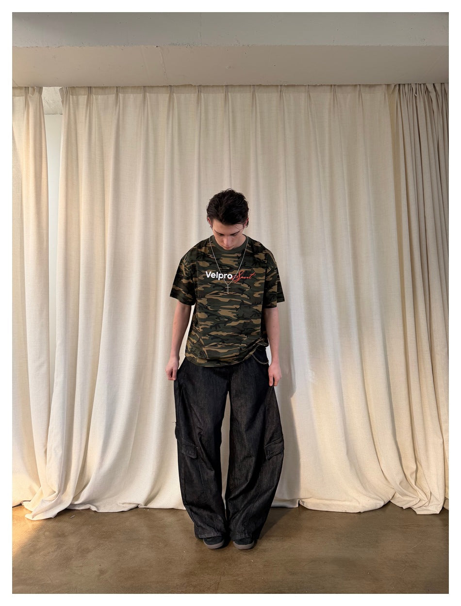 Full-length model looking down in a green and brown camouflage graphic T-shirt and loose black cargo jeans.