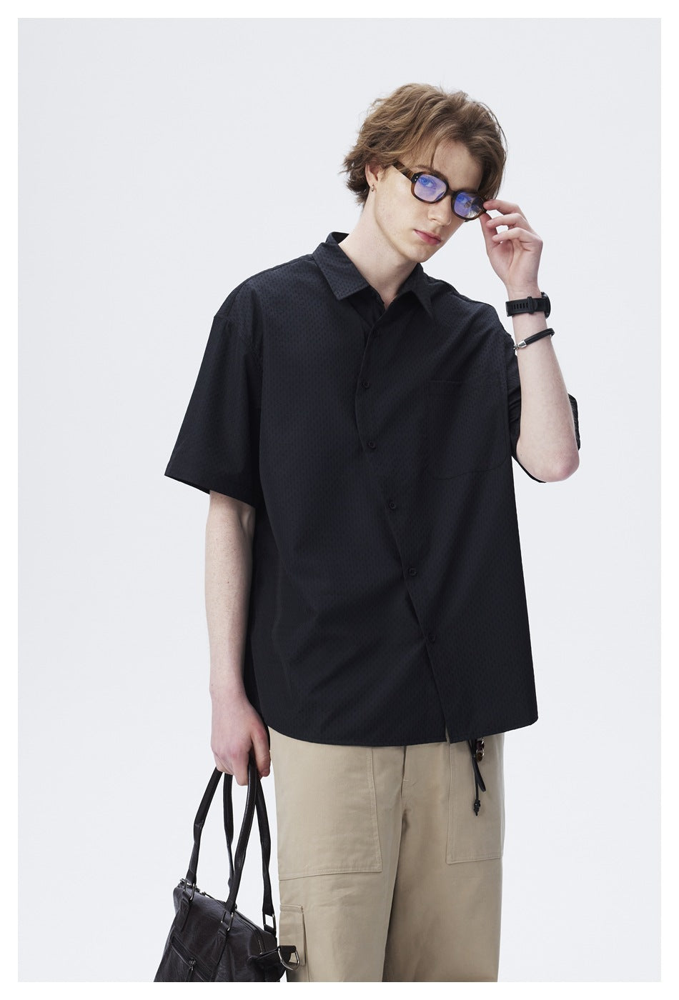 Model wearing a black short-sleeve button-up shirt with a chest pocket and loose beige trousers.