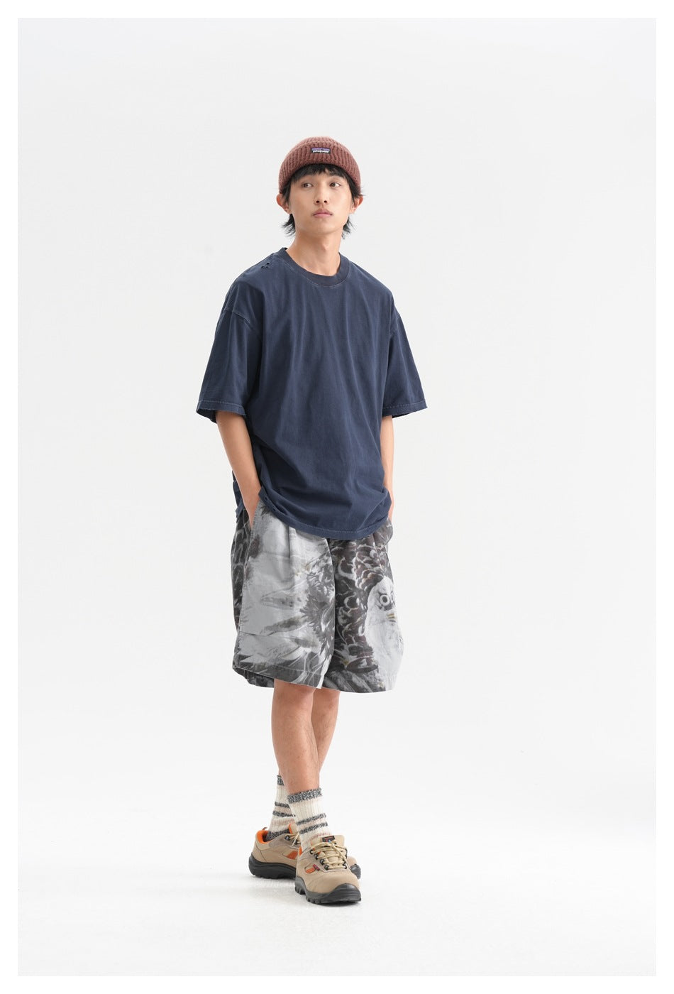 Model in a loose navy T-shirt and gray eagle-print shorts, wearing a rust beanie and hiking shoes.