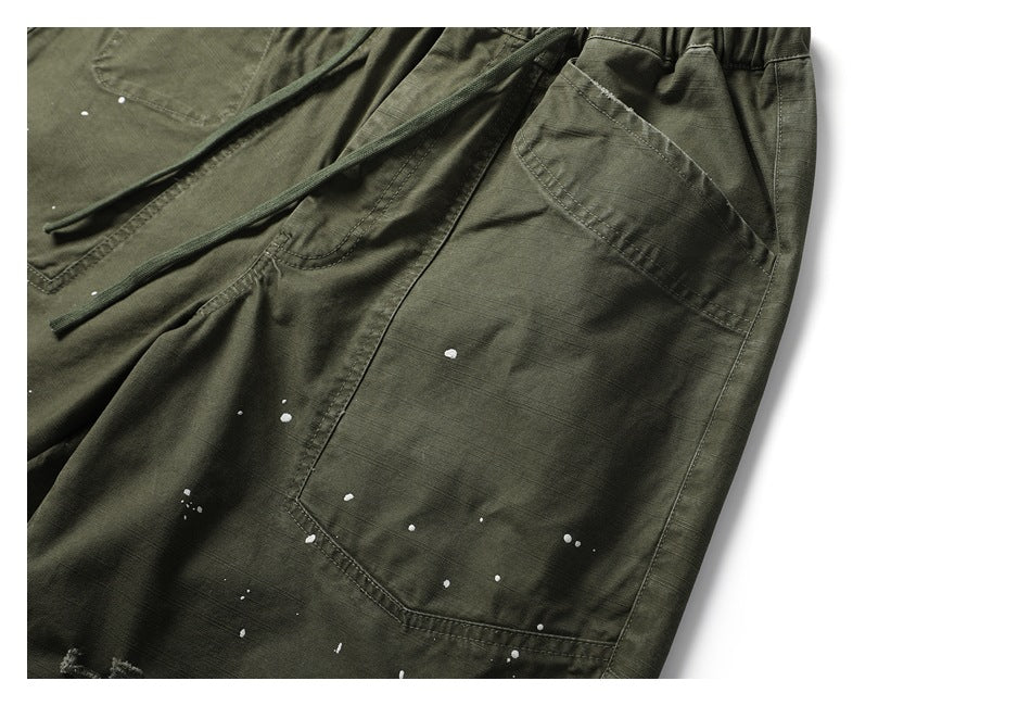 Close-up of an olive-green pant leg with a large stitched pocket and scattered white paint flecks.