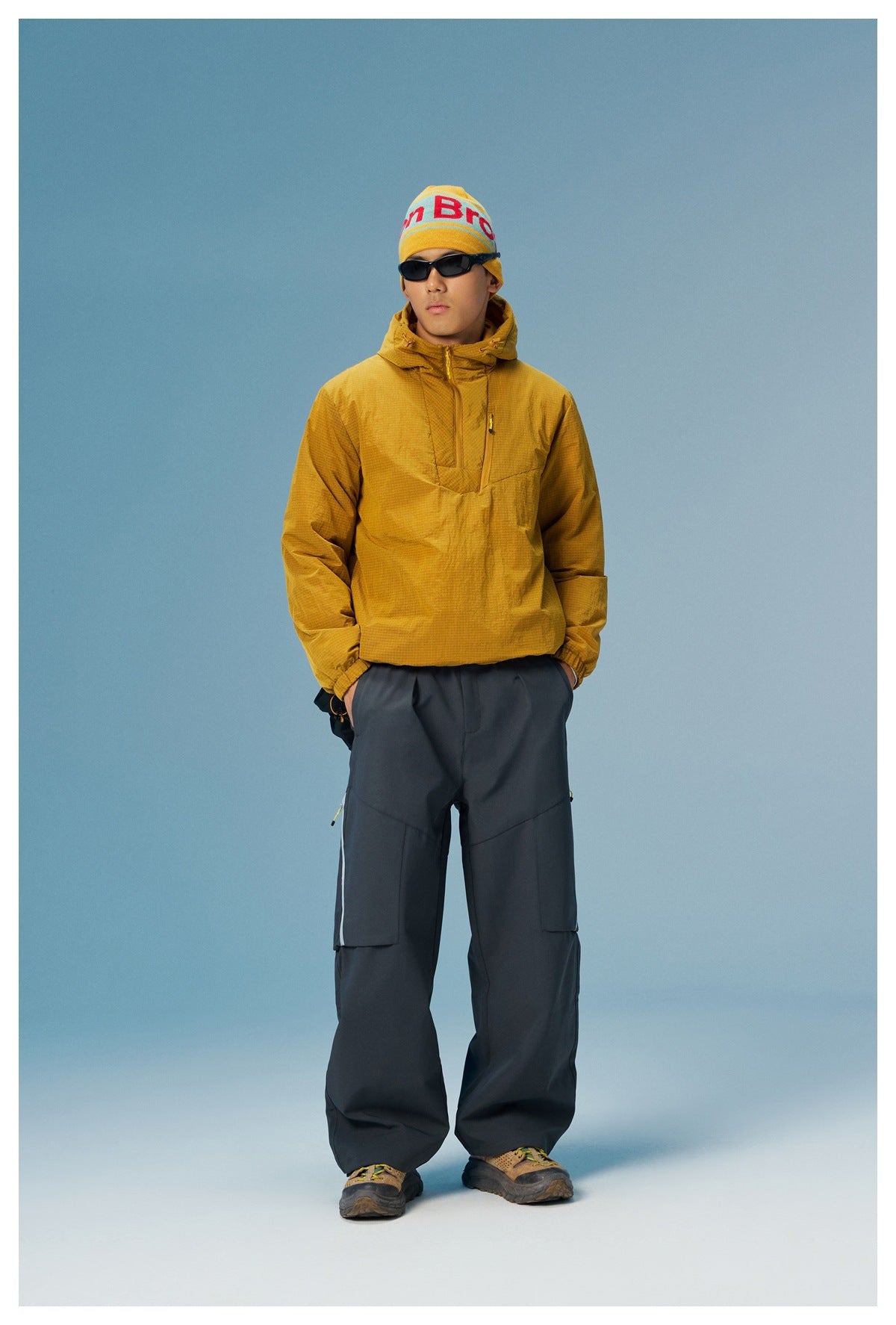 Model wearing a mustard hooded pullover with dark wide-leg pants against a blue studio background.