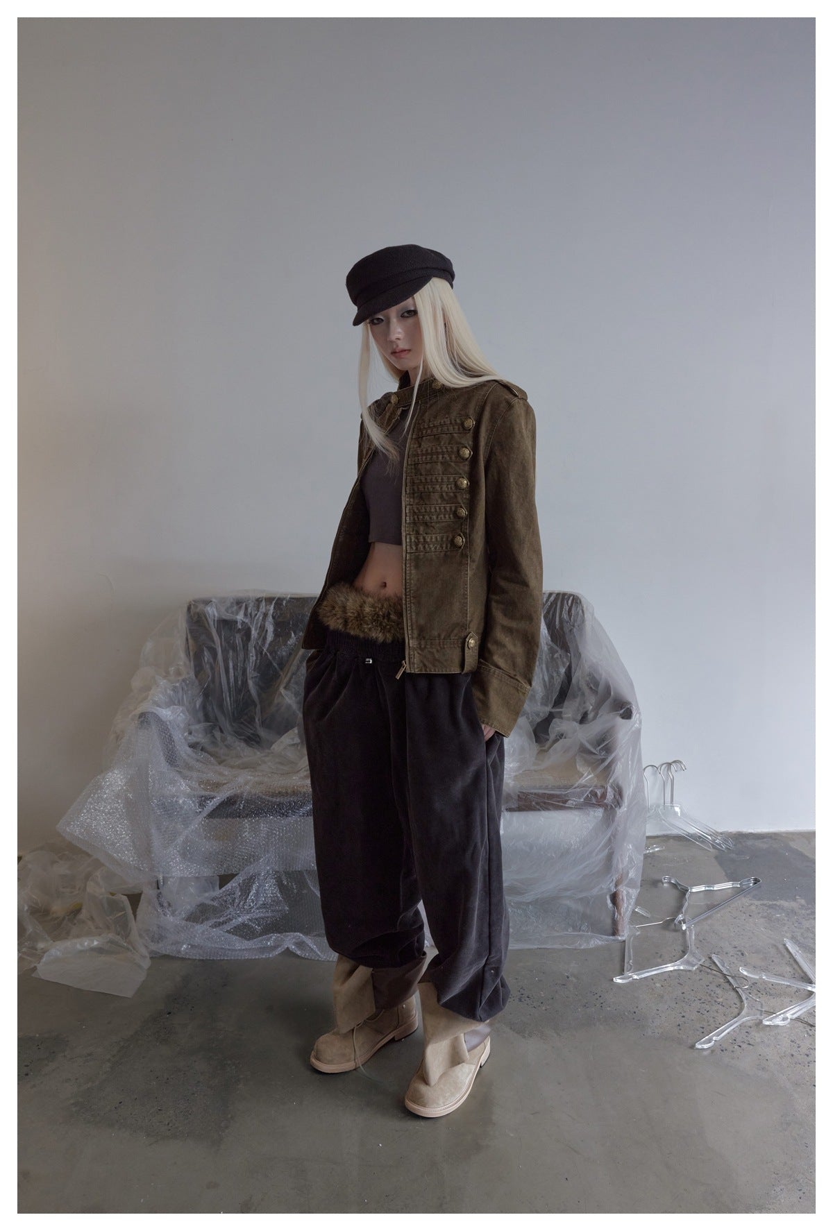 Model in the brown military-style short jacket, angled pose with hand in pocket, dark pants and tan boots