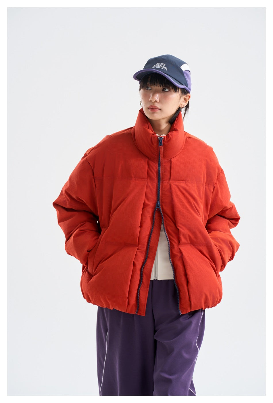 Model wearing an open red-orange puffer jacket with a high collar, purple trousers, and a navy and purple cap.