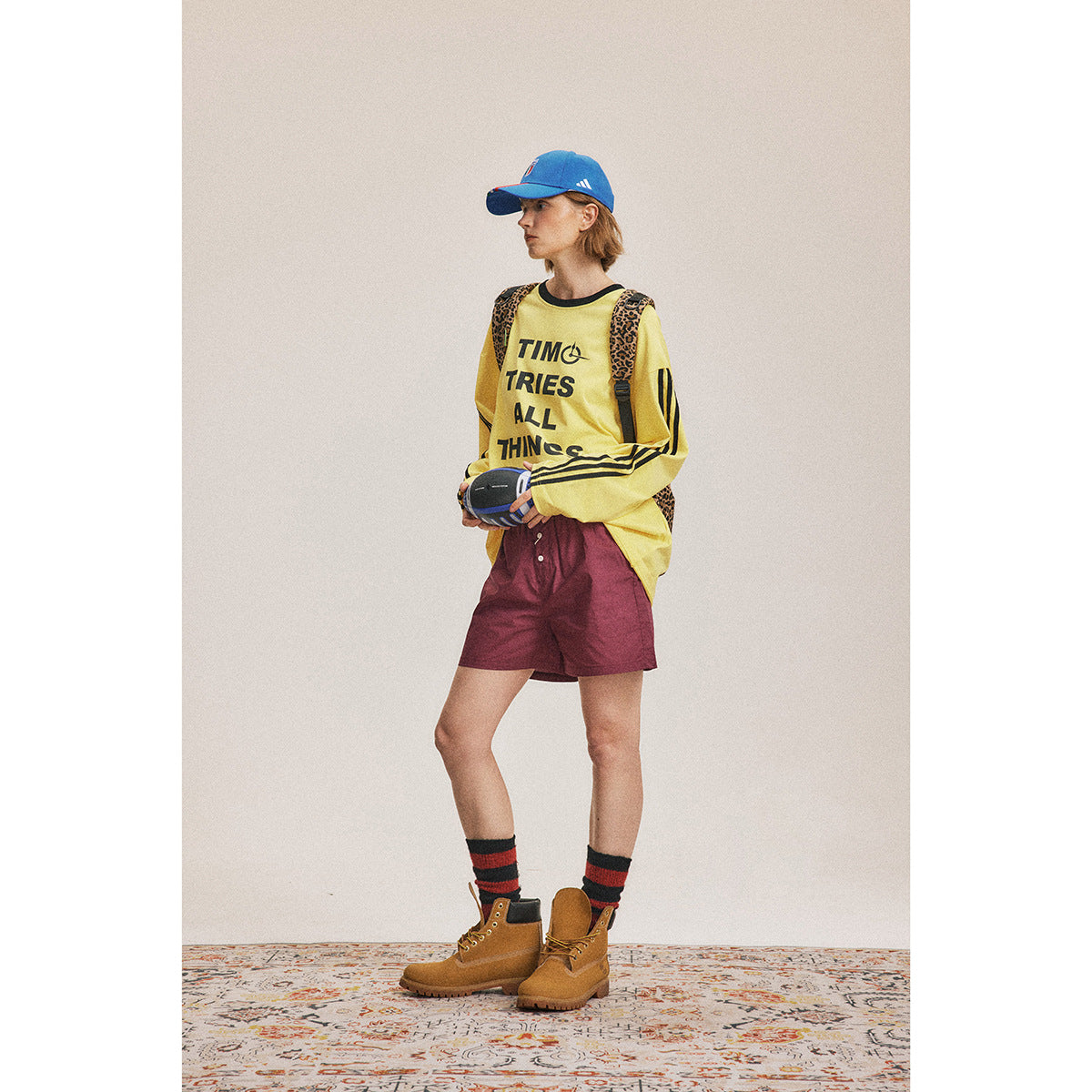 Full-length model wearing a yellow slogan sweatshirt with black sleeve stripes, burgundy shorts, and a leopard-print