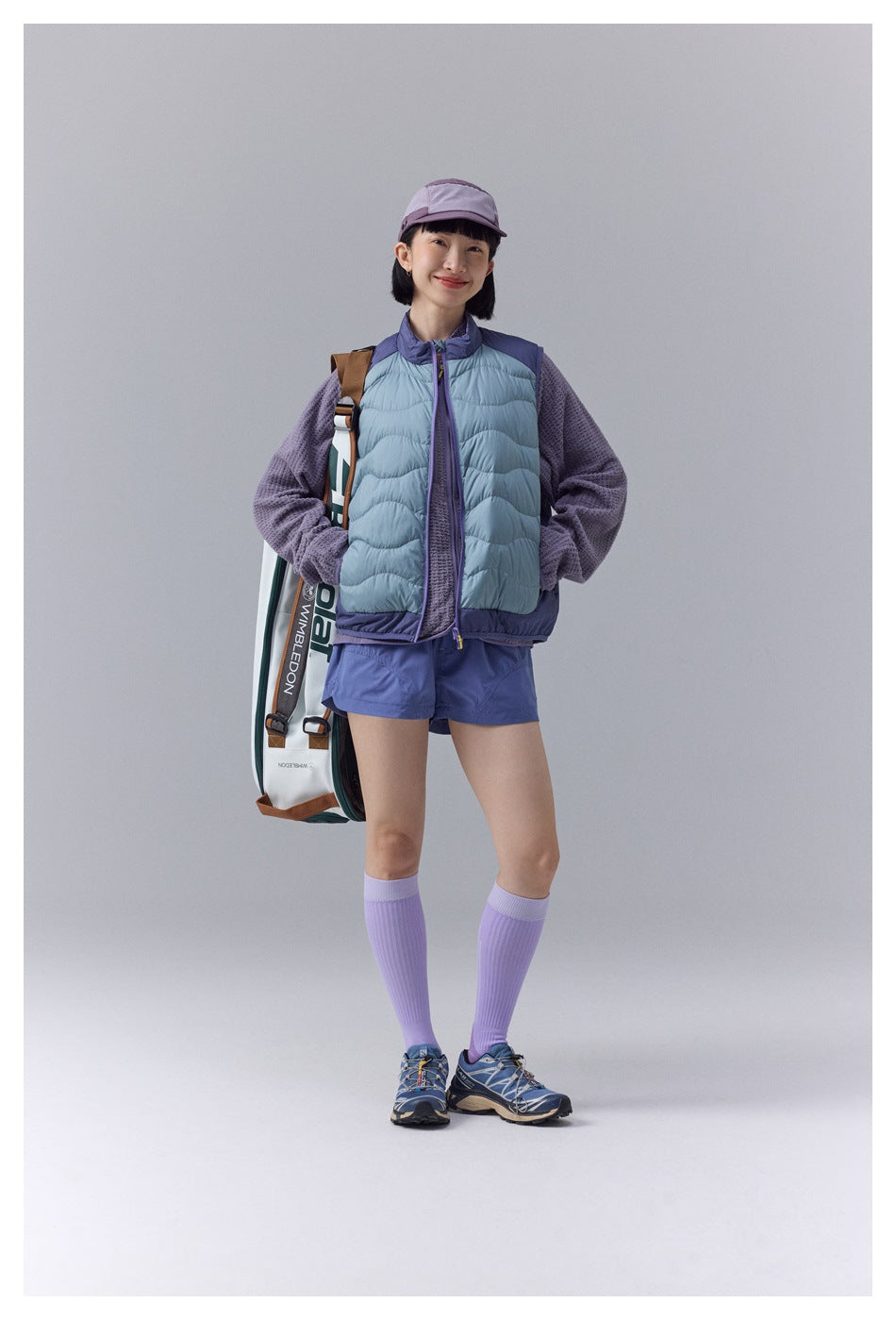 Model wearing a pale blue quilted vest with purple trim over a textured purple top and blue shorts, front view.