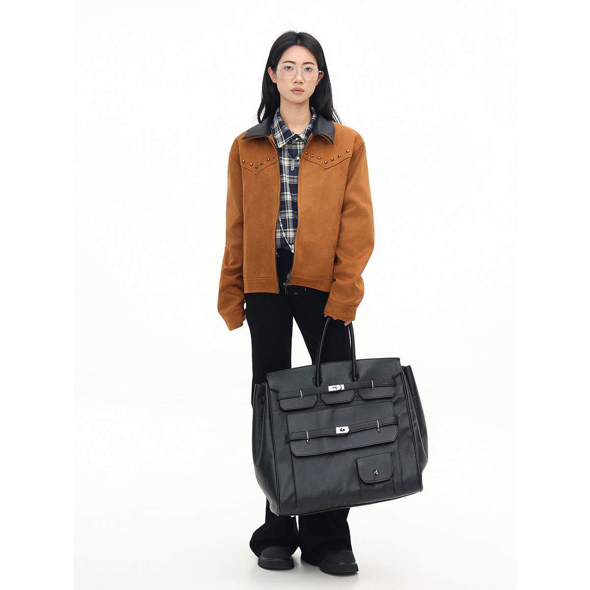 Model wearing a rust-brown studded jacket over a navy plaid shirt and black trousers, carrying a large black tote.