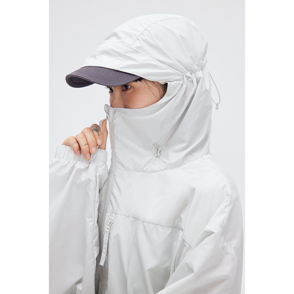 Close-up of a model wearing a white hooded jacket zipped high over the face, with a dark-brimmed cap.