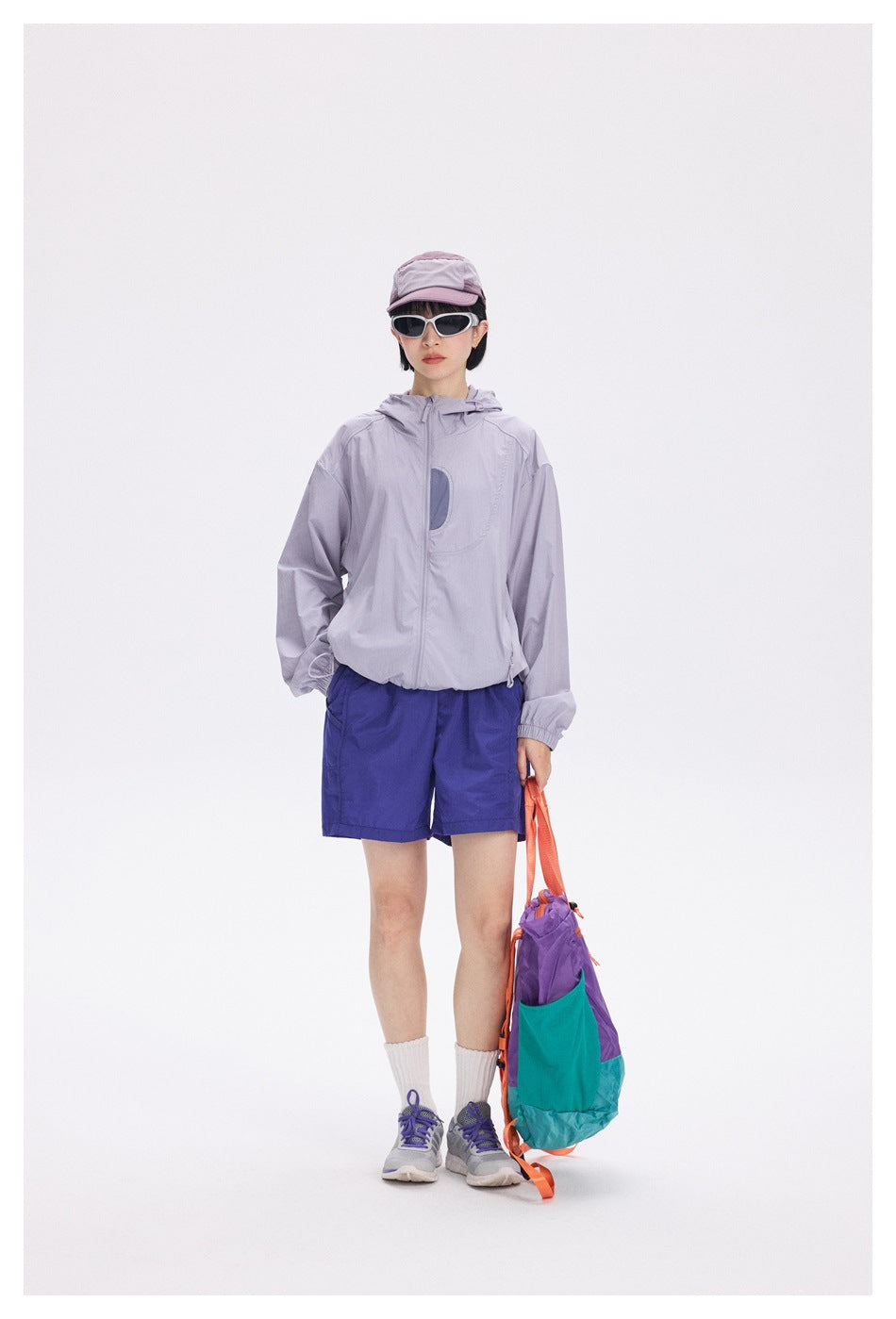 Full-length front view of a model wearing a light lavender hooded jacket, royal blue shorts, a cap and sunglasses.