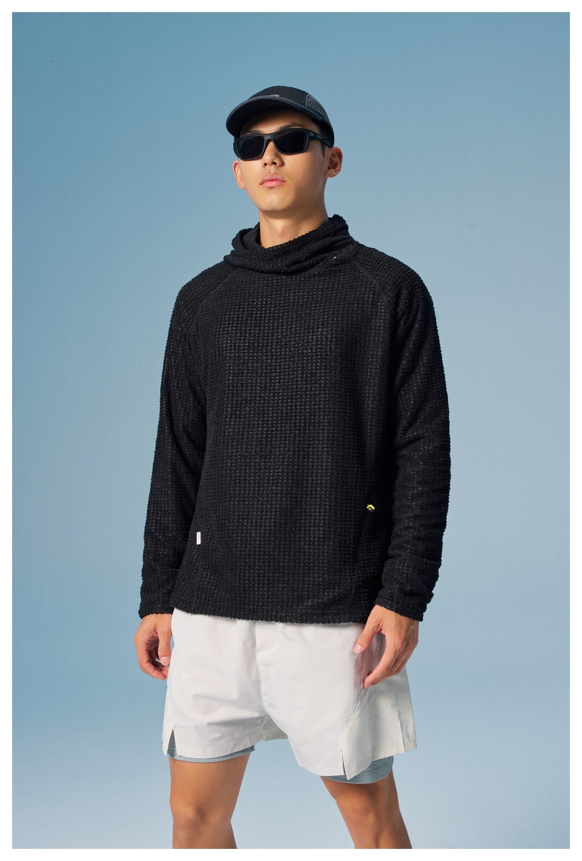 Model wearing a black textured high-neck pullover with white shorts against a blue studio background.