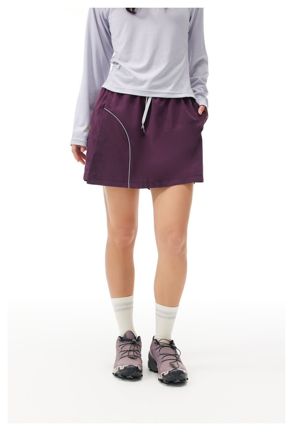 Woman wearing a purple mini skort with curved light piping, a lavender long-sleeve top, white socks and trail shoes