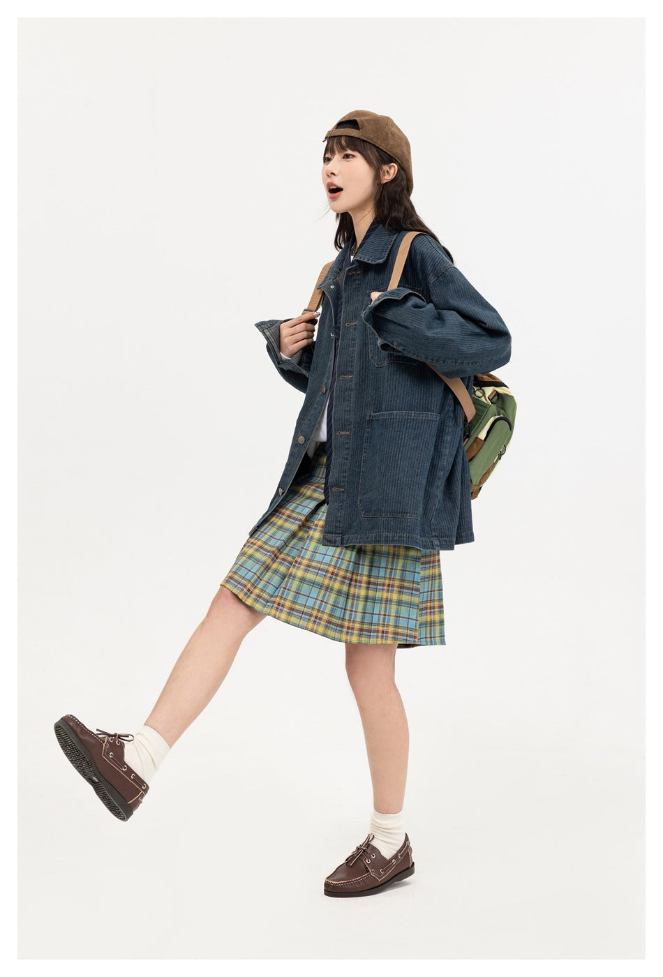 Model in a mid-step pose wearing a dark blue denim jacket with plaid pleated skirt, brown cap, backpack and loafers, side