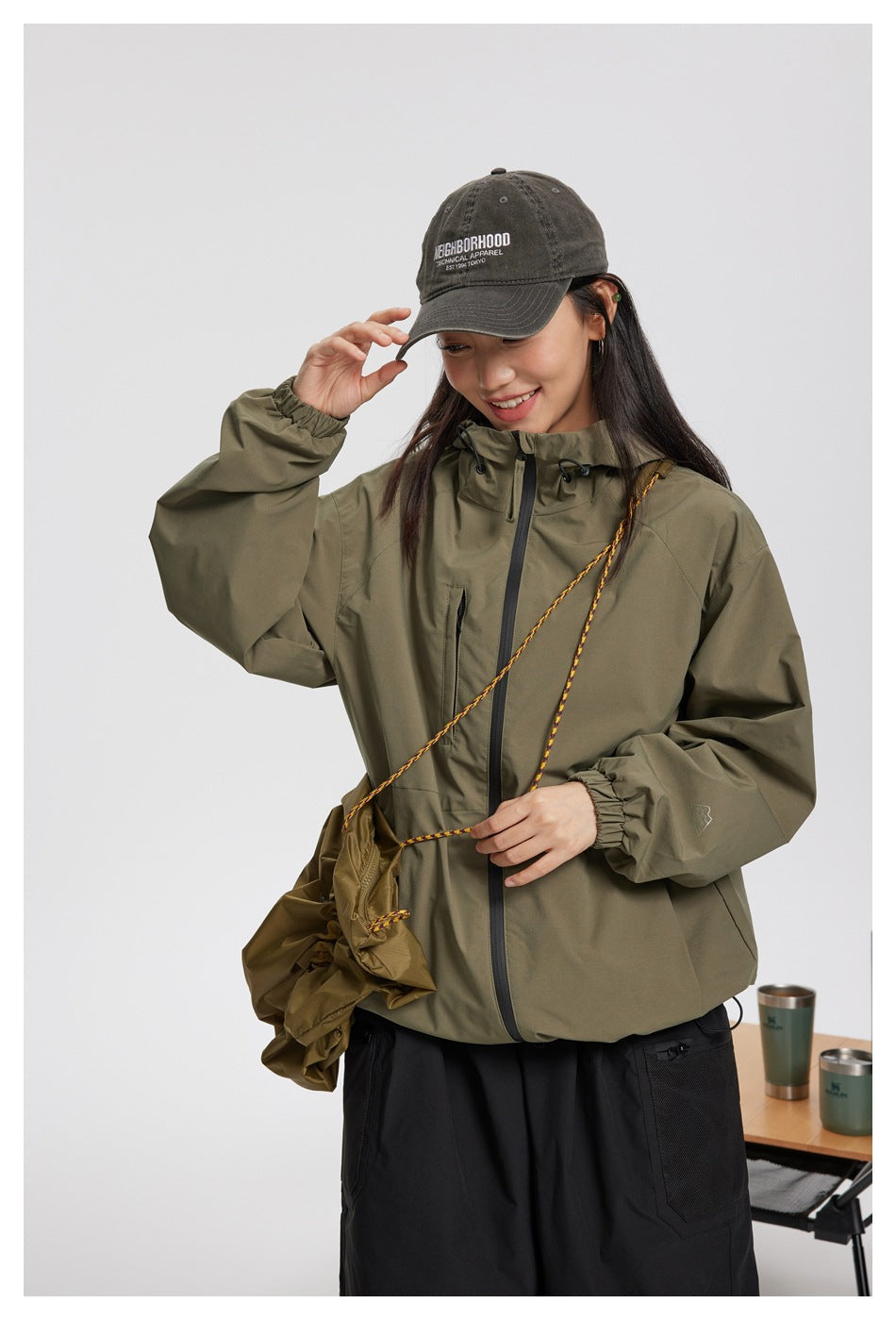 A person models an olive hooded jacket with a front zipper, drawcords, and elastic cuffs.