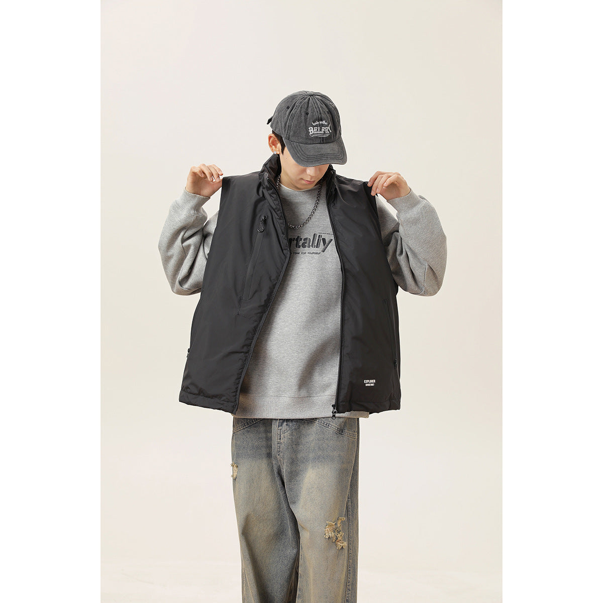Model holds open a black high-collar zip vest, showing its front over a gray sweatshirt.