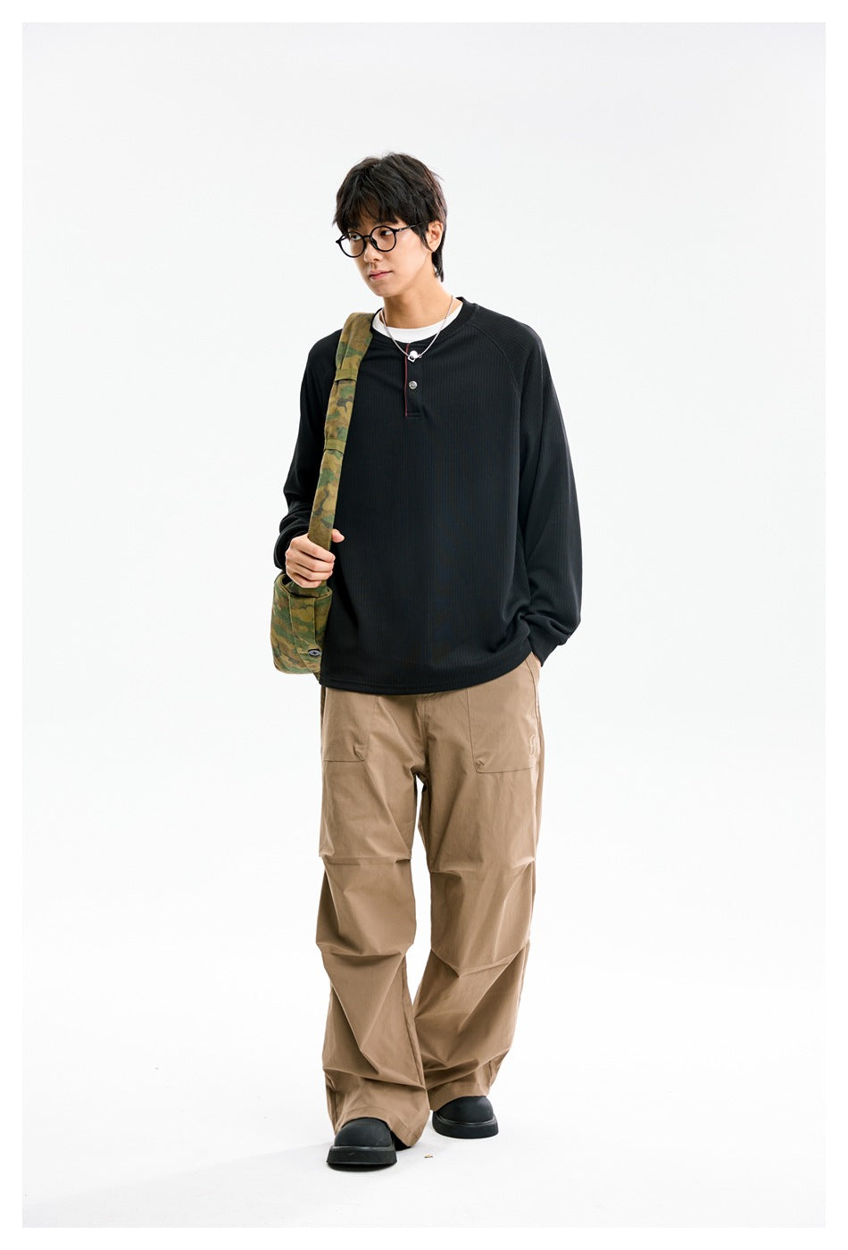 Model wearing a black long-sleeve henley and loose tan pants, carrying a green camouflage-patterned shoulder bag.