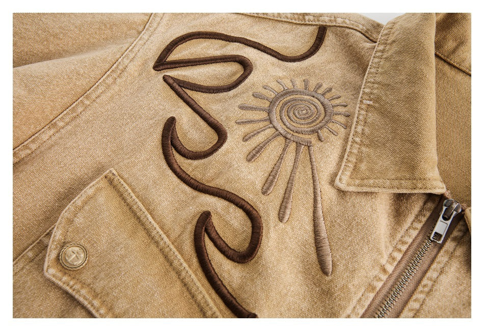 Close-up of a tan jacket with dark brown embroidered curves, a sun motif, collar, and metal zipper.