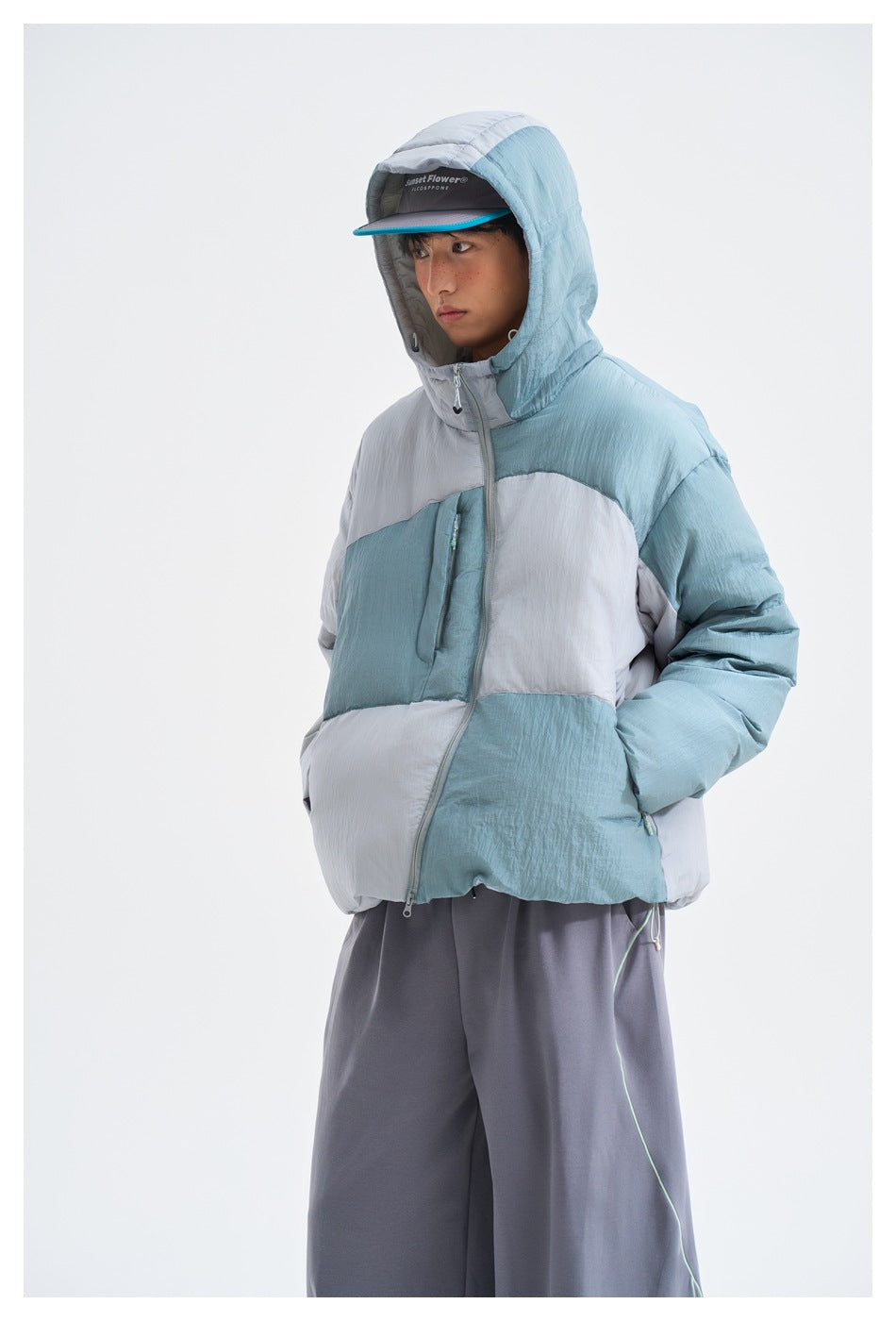 Model wearing a blue-gray color-block hooded puffer jacket with a front zip and matching gray trousers.