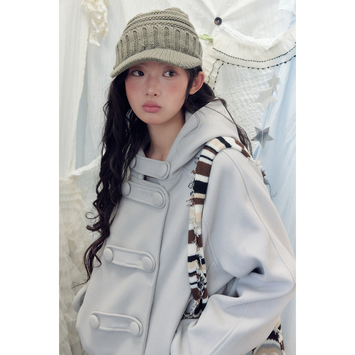 Close view of a model in a pale gray hooded jacket with oversized loop closures and a striped scarf.