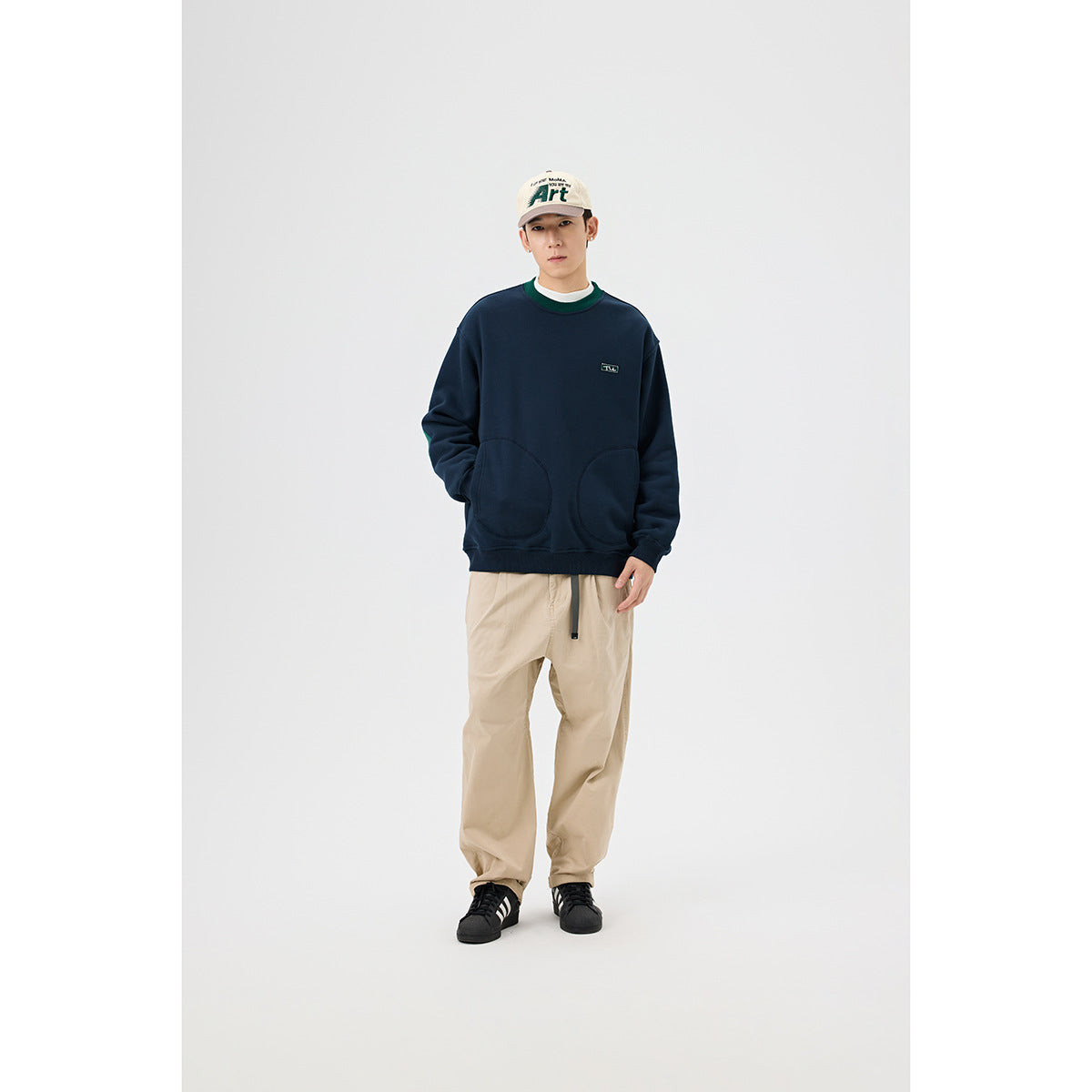 Front view of a model wearing a navy sweatshirt with green collar trim and two rounded front pockets, with beige trousers.