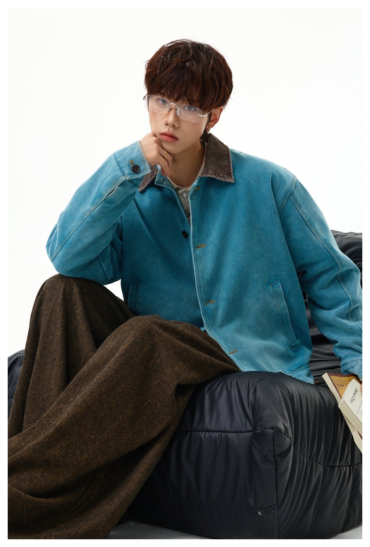 Seated model wearing a teal button-front jacket with a brown collar and dark wide-leg trousers.