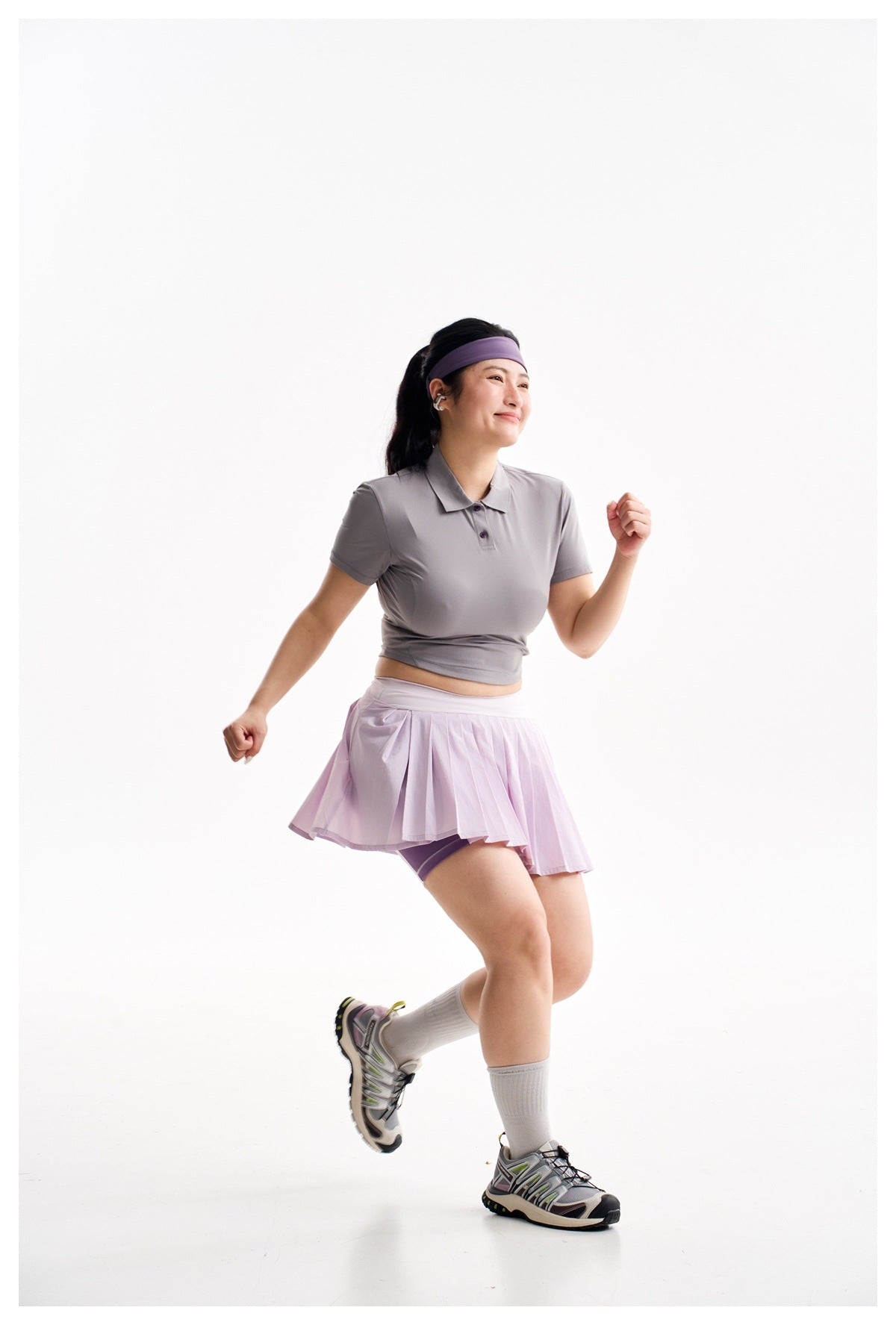 A woman in a gray cropped polo and lilac pleated skort poses in a running stance against a white background.