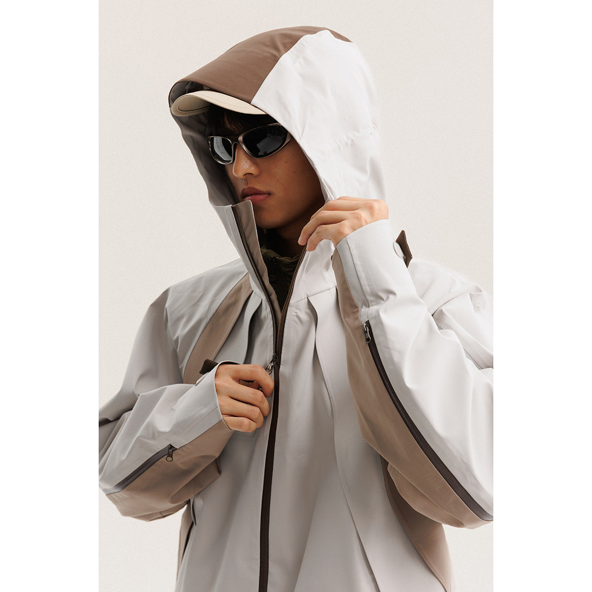 Model wearing a light gray hooded jacket with taupe panels, dark zippers and an oversized hood.