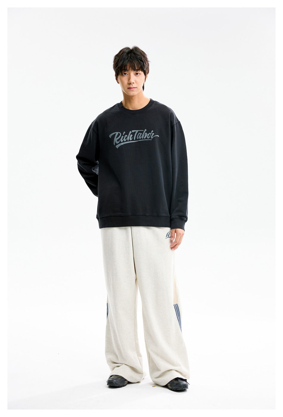 Front view of a model wearing a black script-logo crewneck sweatshirt with loose off-white pants.