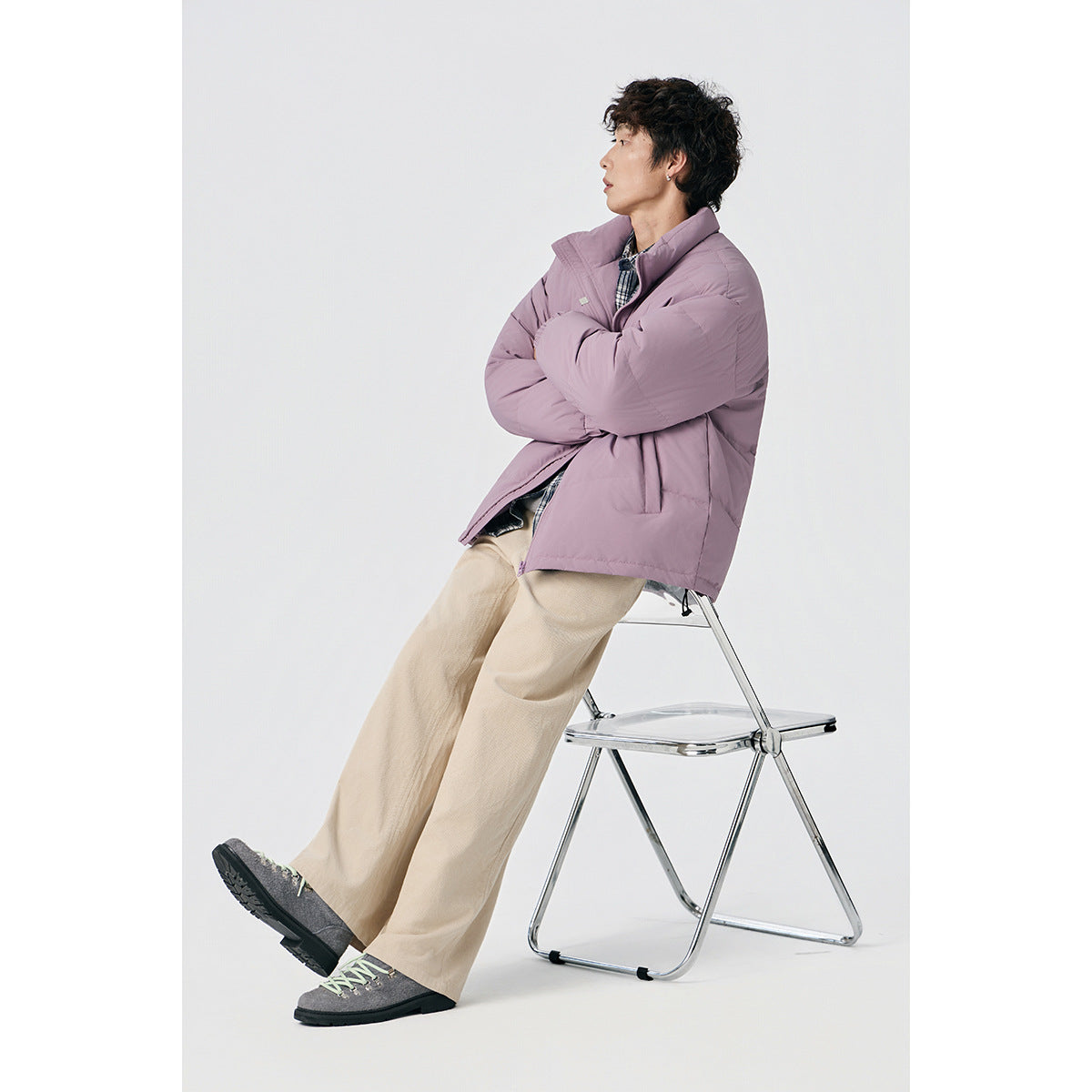 Model seated in a mauve padded jacket with a high collar, crossed arms, and beige wide-leg trousers.