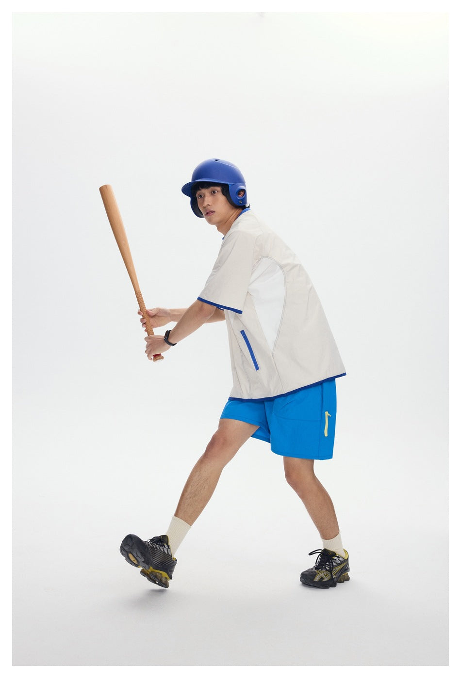 Full-body view of a model in a cream shirt with blue trim and bright blue shorts, holding a bat.