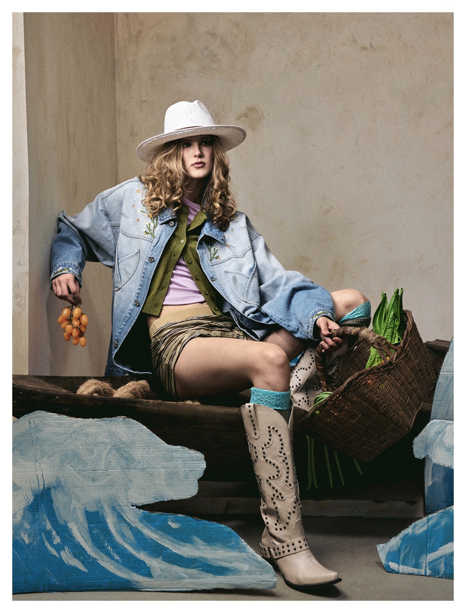 Seated model in an oversized light blue denim jacket with floral embroidery and trim, striped shorts, and tall beige boots.