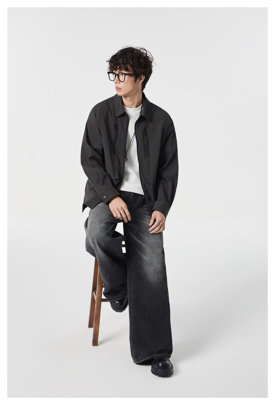 Model seated in a black utility jacket over a white shirt and wide-leg charcoal jeans.