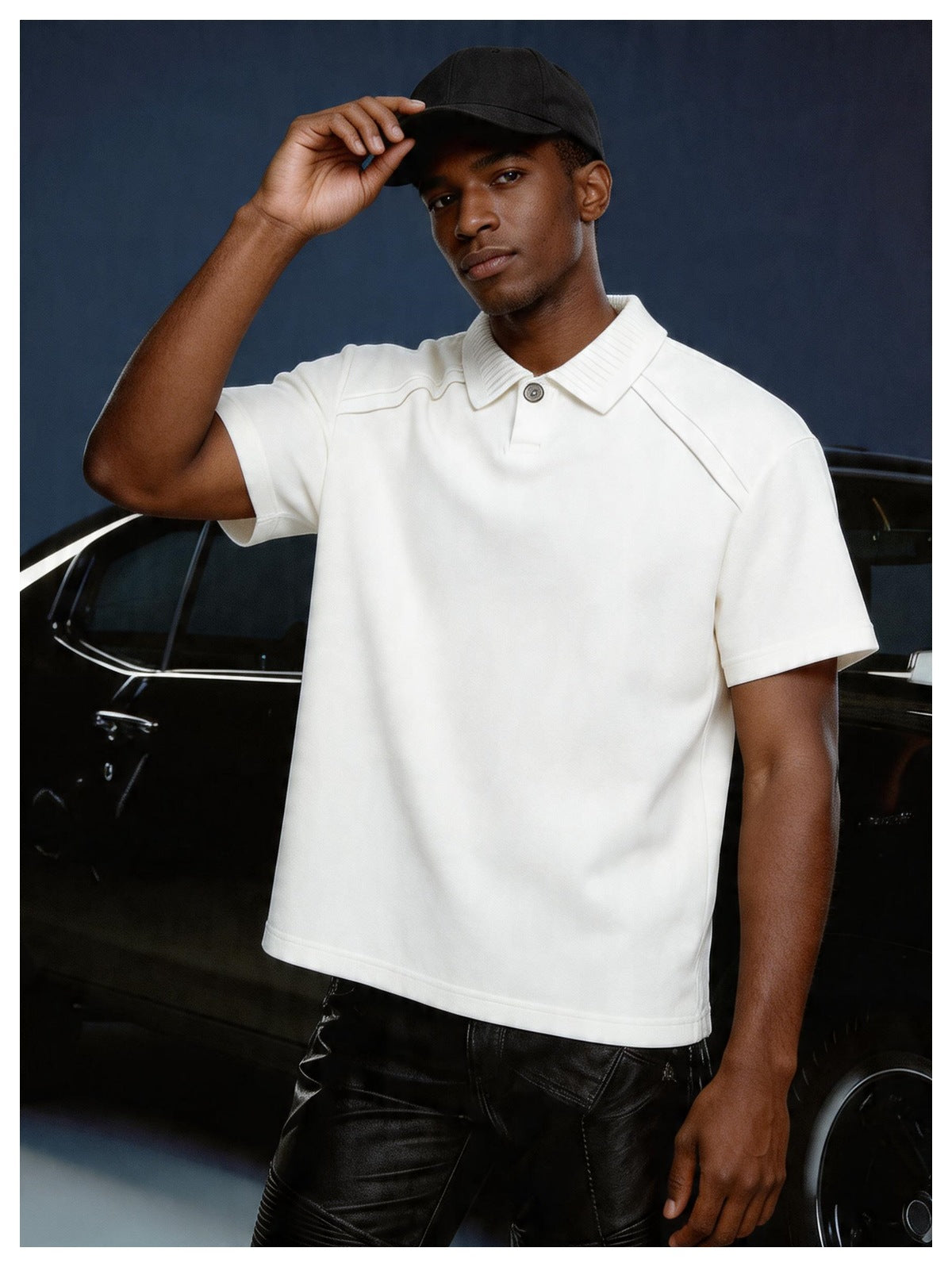 Man in a white short-sleeve polo with ribbed collar and black cap, standing in front of a dark car