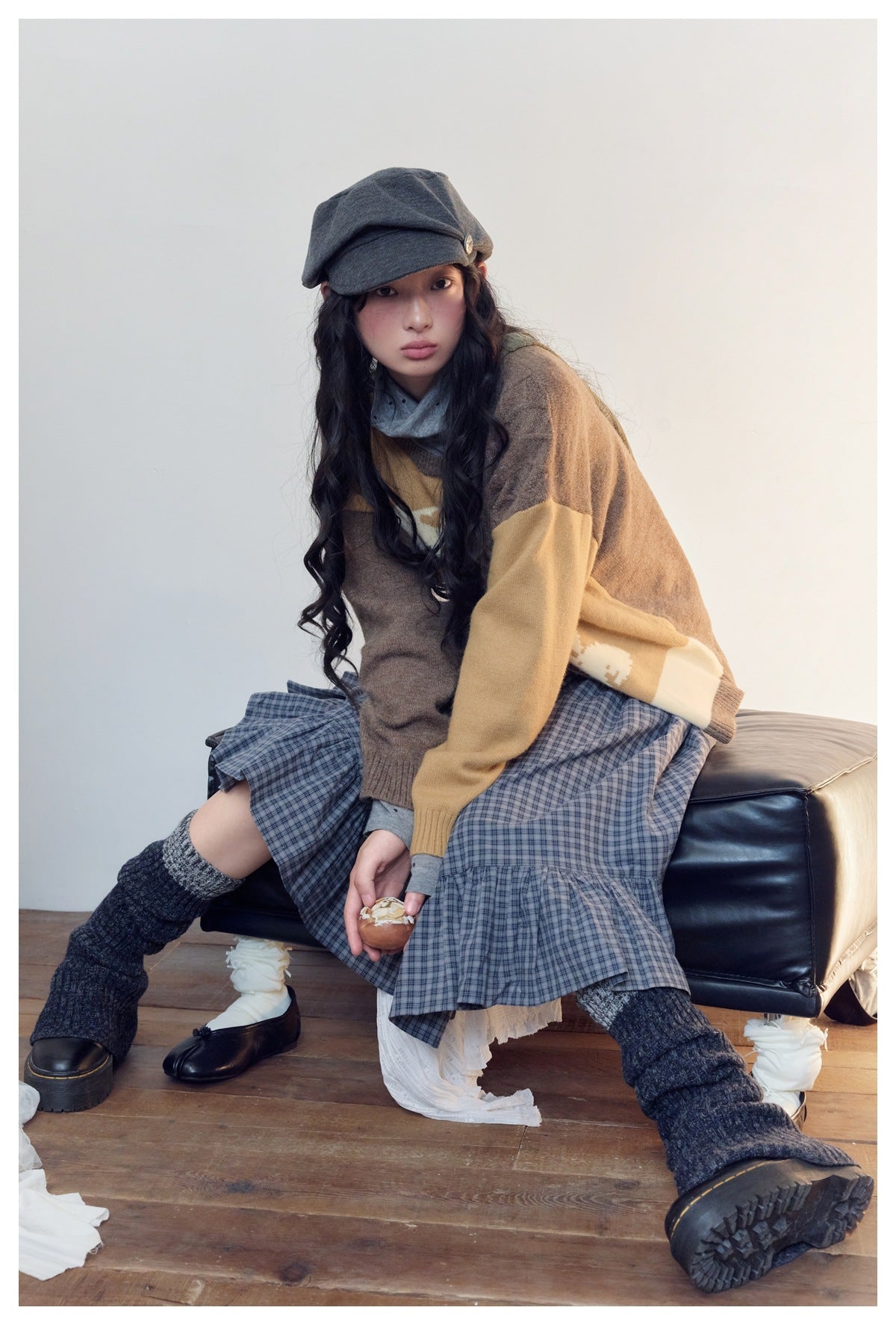 Model sitting in a brown and mustard color-block cardigan and blue plaid skirt, wearing a gray cap.