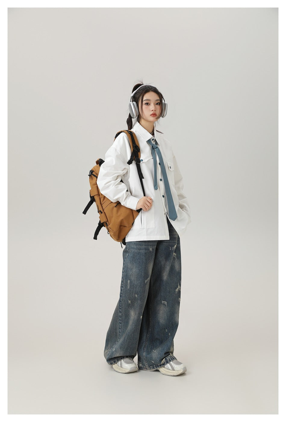 Model in a white oversized shirt with a blue necktie and distressed jeans, carrying a brown backpack and wearing headphones.