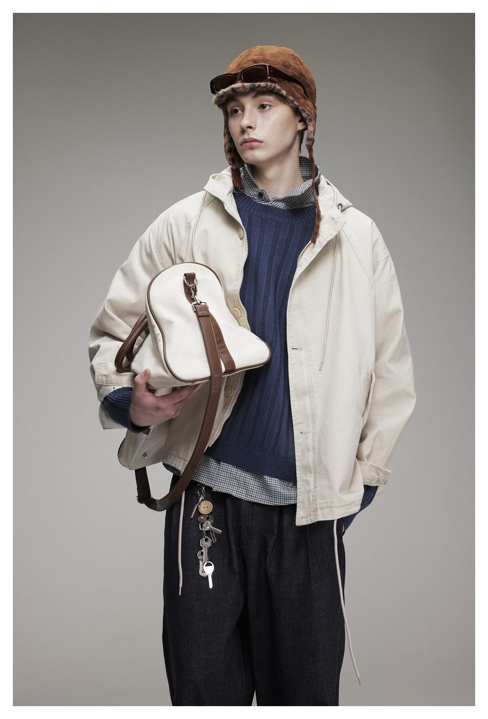 Model in an off-white hooded jacket and navy ribbed sweater, holding a cream bag with brown trim.
