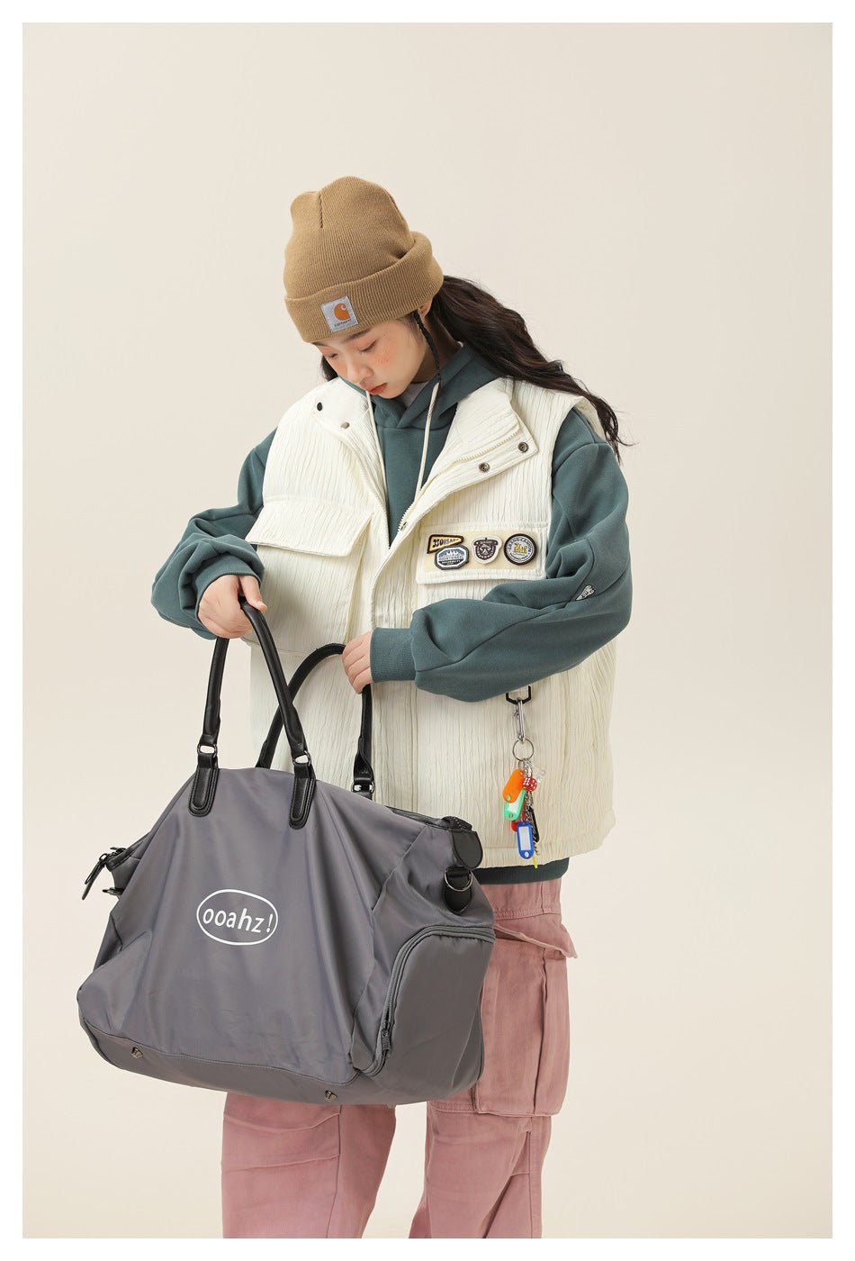 Model wears an ivory utility vest over a teal hoodie with pink cargo pants and holds a gray tote.