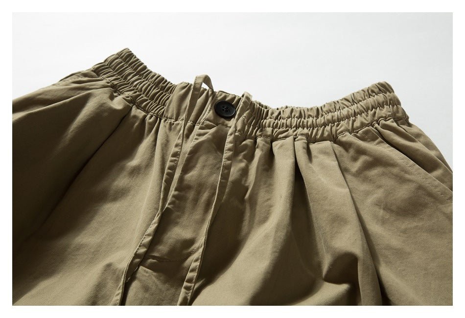 Close-up of khaki trousers showing the gathered elastic waistband, button closure, fly, and front pleats.