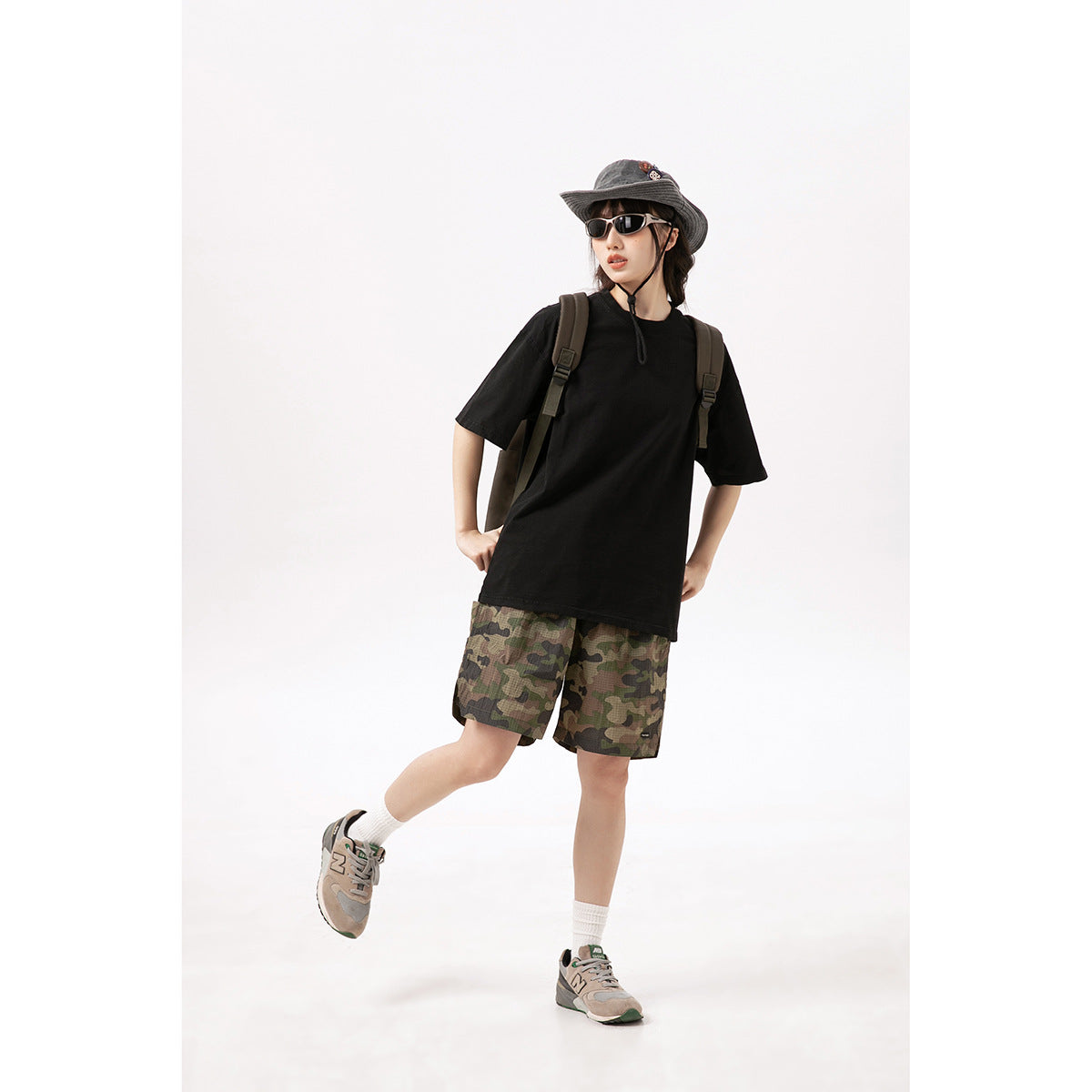 Model wearing camouflage shorts with a black T-shirt, bucket hat, sunglasses, backpack, and gray sneakers.