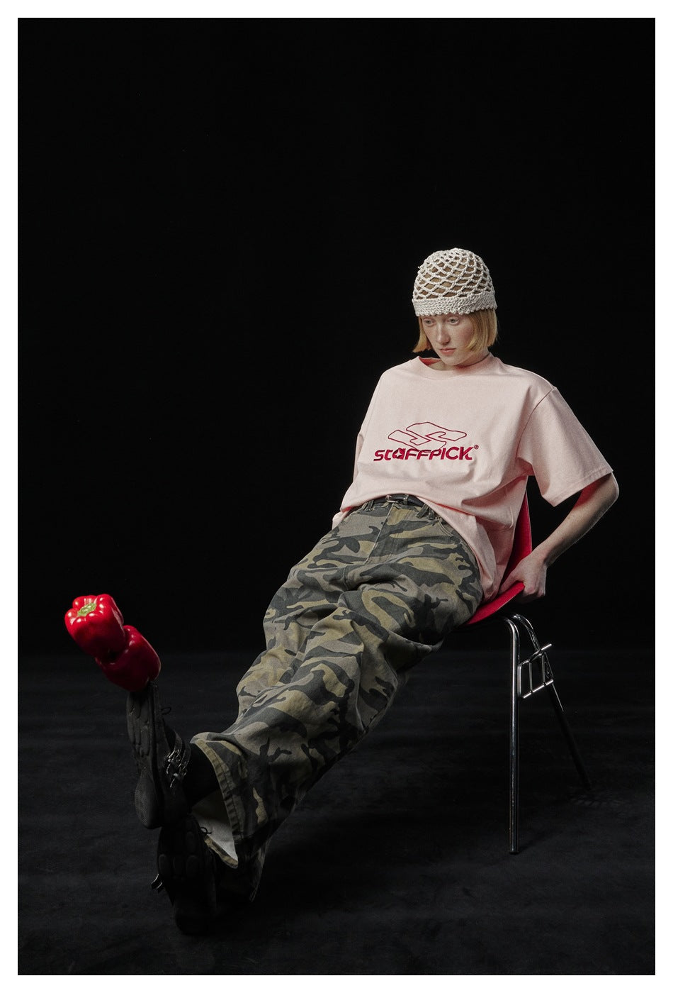Model reclining in a red chair, wearing a pale pink graphic T-shirt and camouflage trousers.
